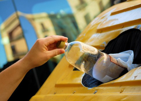 A person with a fair-skinned hand is seen inserting a clear plastic bottle into a large, yellow, rectangular waste disposal unit, which appears to be a recycling or rubbish bin. The bottle has a brown label visible through the transparent plastic and is held upright as it is being placed into the opening of the bin. The yellow bin has a smooth, glossy surface with visible rivets or fasteners around the opening, and appears to be situated outdoors on a driveway or pavement. In the background, blurred building facades and windows suggest an urban or suburban environment, with partially visible blue sky indicating daylight conditions. The scene is professional and neutral, relevant to waste collection or rubbish removal services offered by West Ham House Clearance or similar providers, highlighting the disposal of recyclable bottles in a typical rubbish or recycling bin commonly used in the local area.