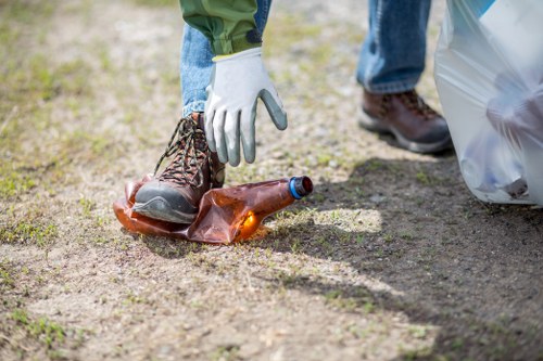 A person wearing blue jeans and white gloves is picking up a discarded brown glass bottle from a patch of dirt and grass on a driveway or outdoor area. The bottle appears to be a used beer or soft drink container, with its cap removed and resting nearby. In the background, there is a transparent plastic rubbish bag, partially filled or tied, positioned on the right side of the image. The ground surface is a mix of soil and small patches of grass, indicating an outdoor setting possibly near a residential property. The scene reflects a clean-up effort, typical of rubbish removal services in West Ham or nearby areas, carried out by West Ham House Clearance to maintain local environments free of litter and debris. The lighting is natural, suggesting daylight, and the overall scene emphasizes environmental cleanliness and professional waste management practices. This image visually supports the importance of rubbish collection, disposal, and site clearance in East London.