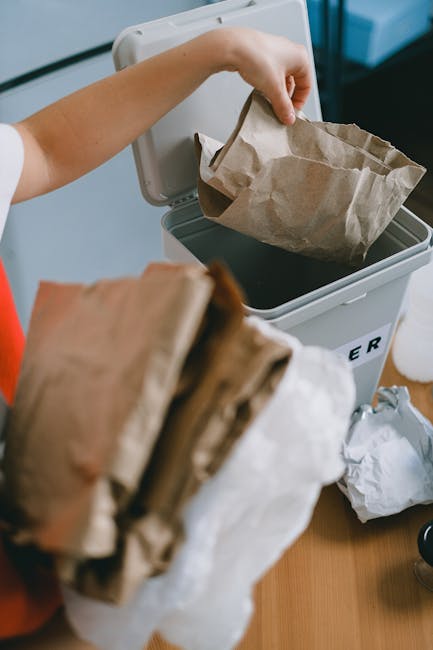 Close-up image of several black plastic rubbish bags tightly tied at the top, with crinkled, shiny surfaces reflecting light, stacked against a plain, light-colored wall. The bags are placed on an out