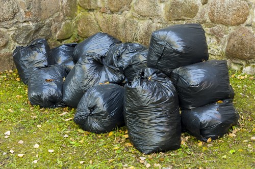 A collection of multiple black plastic rubbish bags, tightly knotted at the top, arranged in a small cluster on a grassy area adjacent to a stone wall made of irregularly shaped stones. The bags appear to contain household waste and are positioned on a patch of green grass with some fallen leaves and small white pebbles scattered around. The stone wall behind them provides a textured, natural backdrop, indicating an outdoor setting, possibly near a residential property in Westham. The scene reflects an area prepared for rubbish collection or disposal, consistent with services offered by Westham House Clearance for local rubbish removal in the Westham area. Natural daylight illuminates the scene, highlighting the glossy finish of the plastic bags and the varied textures of the stone wall and grassy surface.