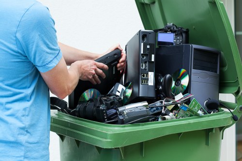 A person wearing a light blue T-shirt is seen lifting a small electronic device from a large green waste bin filled with various discarded electrical items, including desktop computers, computer monitors, DVDs or CDs, and other electronic waste. The electronic devices are stacked and leaning against each other, with some showing visible circuit boards and plastic casings in black, grey, and beige tones, with metallic and reflective surfaces on the discs. The bin is situated outdoors on a paved surface, possibly in a residential or commercial area in Westham, with natural daylight illuminating the scene. The image emphasizes the process of rubbish removal and electronic waste disposal, consistent with services provided by Westham House Clearance in the local area.