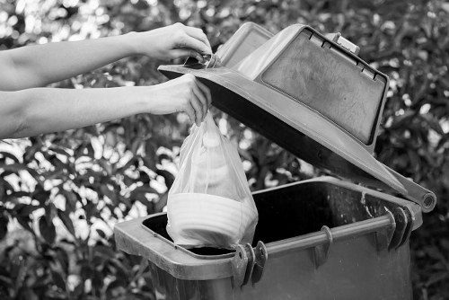 A person is opening the lid of a large outdoor rubbish bin, revealing a black interior with a partially filled plastic bag containing multiple white bowls or dishes. The person's arms and hands are visible, wearing short sleeves, and they are in the process of either placing items into or removing items from the bin. The bin is situated on a paved surface with dense foliage and bushes in the background, suggesting a residential garden or driveway setting. The scene appears to be during daylight, providing natural lighting that highlights the textured surfaces of the bin, the plastic bag, and the bowls inside. This image relates to rubbish removal or waste disposal services in Westham and reflects routine household rubbish management tasks, typical of waste clearance operations undertaken by companies like Westham House Clearance in the area.