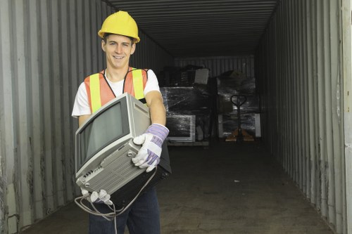 A young man wearing a yellow safety helmet, high-visibility vest with orange and yellow reflective strips, white T-shirt, and gloves is standing at the entrance of a storage unit or container on a driveway. He is holding an older-model computer monitor, which appears to be in good condition, with his left hand gripping the top and his right supporting the bottom. The background inside the container shows several large black plastic wrapping or tarpaulin covers, alongside other indistinct items stacked behind. The container has corrugated metal walls and a dark interior, with natural daylight illuminating the scene. The surrounding environment suggests a residential or commercial property in Westham, possibly in East Sussex, where Westham House Clearance operates. This scene indicates a scene of rubbish clearance, with the worker preparing to remove electronic waste as part of a professional house clearance service.